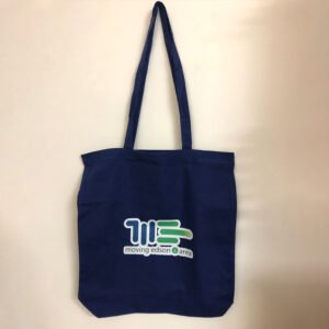 The Moving Edson Tote Bag is navy blue with two handles and features the Moving Edson Forward logo in white, green, and blue on the front. It is displayed flat on a light-colored surface.